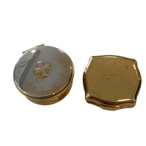 Könnte beinhalten: Zwei Vintage-Kompaktpuder. Eines ist rund mit silbernem Deckel, goldenem Rand und goldenem Kreuz. Das andere ist ein goldfarbenes, achteckiges Kompaktpuder mit Webmuster und dem Namen "Stratton" auf der Vorderseite.