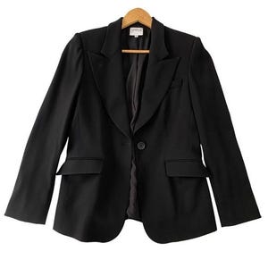 Puede incluir: Una blazer negra con solapa de muesca, dos bolsillos delanteros y cierre de un solo botón. La blazer está colgada de una percha de madera. La etiqueta de la blazer dice "Armani".