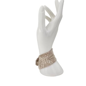 Peut inclure: Un présentoir à bijoux en céramique blanche en forme de main tenant un bracelet large et texturé. Le bracelet est de couleur or clair et présente un motif répétitif de petits éléments carrés.