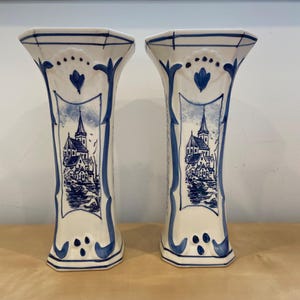 Peut inclure: Deux vases en céramique blanche assortis avec un motif bleu et blanc de Delft néerlandais. Chaque vase présente une illustration détaillée d'une scène de village avec une église et des maisons. Les vases ont un dessus évasé et une base géométrique.