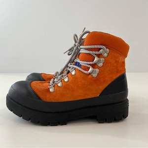 Puede incluir: Botas de senderismo naranjas con suelas y punteras negras. Las botas tienen ojales metálicos plateados y cordones grises y blancos. La parte superior de la bota es de ante naranja, y el cuello es de un naranja más oscuro. Para actividades al aire libre.