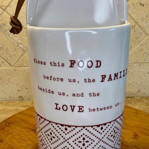 May include: White ceramic container with a brown leather strap. The container has the words "Bless this FOOD before us, the FAMILY beside us, and the LOVE between us" in a dark red font. A geometric pattern decorates the bottom.