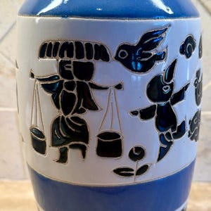 May include: A blue and white ceramic vase with a black and white design of people and birds. The vase has a blue glaze and a white band with a black and white design around the middle.