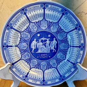 May include: A decorative blue and white ceramic plate featuring a calendar for the year 1997. The plate has a circular design with months labeled around the edge and illustrations in the center. It is displayed on a white wooden stand.