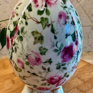 May include: A white ceramic egg-shaped vase with a floral pattern of pink roses and green leaves. The vase has a gold trim around the top and bottom and a gold-trimmed base.