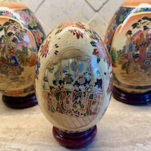 May include: Three decorative ceramic eggs with intricate hand-painted designs of people and flowers. The eggs are on wooden stands.