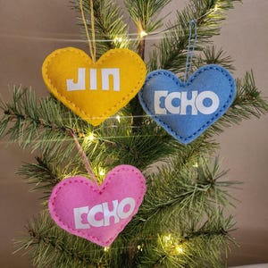 May include: Three heart-shaped Christmas ornaments in yellow, blue, and pink, each with white text. The yellow ornament says "JIN", while the blue and pink ornaments say "ECHO". They hang from a Christmas tree with lights.