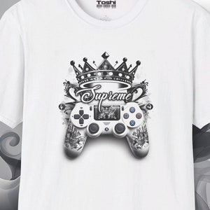Op de afbeelding: Wit T-shirt met een zwart-wit grafisch ontwerp van een videogamecontroller met een kroon en het woord "Supreme" erboven.