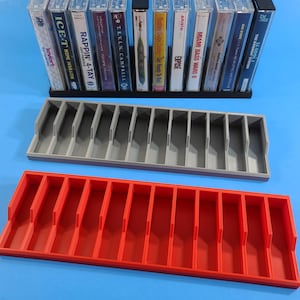 May include: Two cassette tape storage organizers, one gray and one red, with 12 slots each. The organizers are on a blue surface. The gray organizer is in the background and the red organizer is in the foreground.