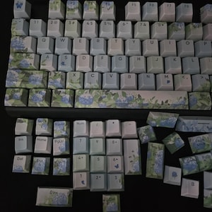 May include: A blue and white keyboard with floral keycaps. The keyboard has a standard layout with function keys, number keys, and arrow keys. The keycaps are decorated with a floral pattern, including hydrangeas and leaves.