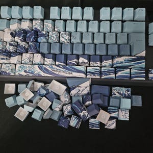 Pode incluir: Um teclado mecânico com teclas personalizadas. As teclas apresentam um padrão de ondas em tons de azul e branco. Algumas teclas são azul claro, enquanto outras apresentam desenhos de montanhas e ondas. Uma pilha de teclas soltas está na parte inferior.