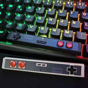 May include: A black and white NES style controller with two red buttons and a directional pad. The controller is sitting on top of a black keyboard with colorful keys.