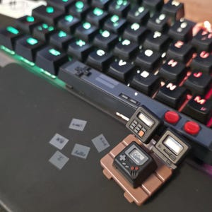 Könnte beinhalten: Eine Sammlung von elektronischen Miniaturgeräten, darunter eine tragbare Spielkonsole, ein Taschenrechner und ein Computermonitor, die auf einer braunen Plattform neben einer schwarzen mechanischen Tastatur mit beleuchteten Tasten liegen.