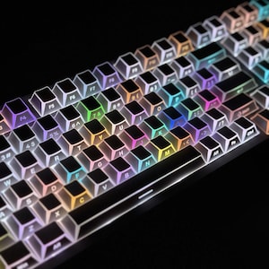 Könnte beinhalten: Eine Computertastatur mit schwarzen Tasten und weißen, durchscheinenden Rändern, durch die eine lebendige, mehrfarbige Regenbogen-LED-Hintergrundbeleuchtung scheint. Das Layout umfasst Standard-alphanumerische Zeichen, Funktionstasten und eine Leertaste.