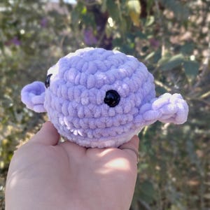 May include: A small, crocheted plush toy shaped like a purple octopus with black eyes. The octopus is sitting in the palm of a hand.