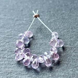 May include: A strand of pale pink and purple gemstone beads strung on a silver wire. The beads are faceted and have a teardrop shape.