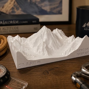 Puede incluir: Modelo blanco impreso en 3D de una cordillera, con el texto "K2 8.611 m" y coordenadas. El modelo está sobre una superficie de madera, con una brújula, una cuerda y una taza con "ADVENTURE".