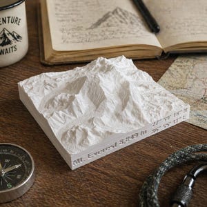 May include: A white, textured 3D model of Mount Everest, with coordinates and elevation details. A compass, a mug with "Adventure Awaits" text, and a map are also visible, suggesting a travel or adventure theme.