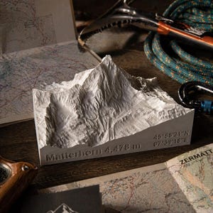 以下が含まれることがあります： 「Matterhorn 4,478 m」の文字と座標が刻まれた白いマッターホルンの山岳模型。木製の表面に、登山用具と地図が配置されています。模型の高さは約448cmです。