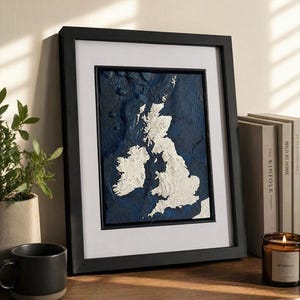May include: A framed relief map of the British Isles, rendered in white against a dark blue backdrop. The artwork is encased in a black frame with a white mount. The piece is displayed on a wooden surface, alongside a mug, plant, and books.
