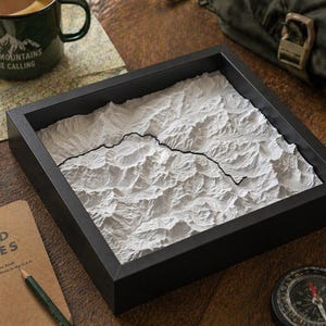 May include: A framed, three-dimensional white mountain relief map with a black border. The map is set in a black frame and sits on a wooden surface. A green mug with the text "The Mountains Are Calling" is visible.