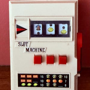 May include: A small, off-white plastic slot machine toy featuring three spinning reels displaying a bell and two lemons. The front includes the text SLOT MACHINE, three red buttons, a red side lever, and a payout chart with various symbols.