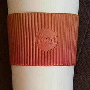May include: A white travel mug with a matching lid and a ribbed, burnt orange silicone sleeve. The sleeve features a circular embossed logo in the centre that reads Food Network in lowercase letters. The cup has a tapered cylindrical shape.