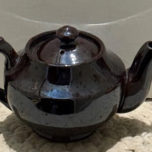 May include: A small, dark brown ceramic teapot with a glossy finish. The teapot has a curved handle, a spout, and a lid with a small knob. The teapot is sitting on a textured surface.