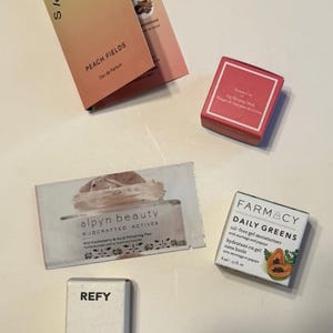 May include: An assortment of beauty products, including a perfume sample, lip sleeping mask, moisturizer, and gloss highlighter. The perfume sample is labeled "Peach Fields" and the highlighter is 0.17 US fl oz.