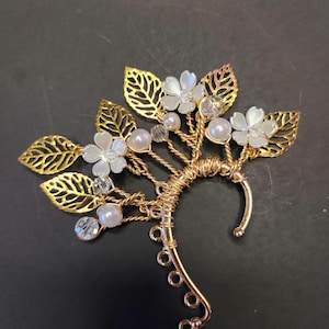 May include: A gold-colored ear cuff featuring a floral and leaf design. The cuff has gold filigree leaves, white flowers, faux pearls, and clear crystal accents. The ear cuff is designed to be worn on the ear.