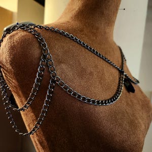 May include: A gunmetal-colored chain shoulder piece with multiple strands and a draped design. The jewelry is displayed on a brown mannequin, showcasing the intricate chain links and overall style. The piece appears to be handmade.