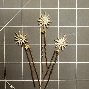 May include: Three gold hair pins with a starburst design featuring clear rhinestones.