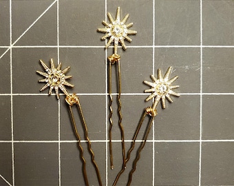 May include: Three gold hair pins with a starburst design featuring clear rhinestones.