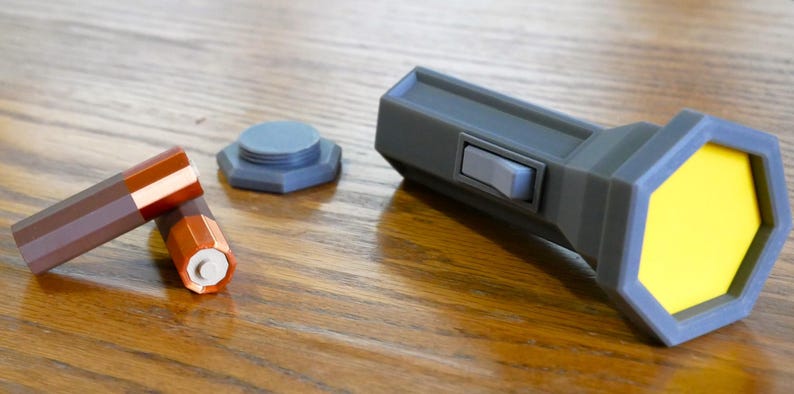 Puede incluir: Una linterna gris con una lente amarilla y un bot&oacute;n negro. Tambi&eacute;n se muestran dos bater&iacute;as marrones y una tapa de bater&iacute;a gris.