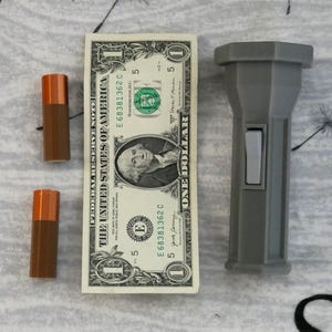 Puede incluir: Una linterna gris con un interruptor negro. La linterna est&aacute; al lado de un billete de cinco d&oacute;lares con el texto "THE UNITED STATES OF AMERICA" y "ONE DOLLAR".