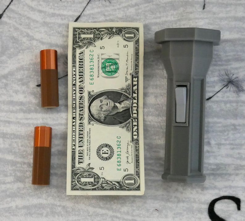 Puede incluir: Una linterna gris con un interruptor negro. La linterna est&aacute; al lado de un billete de cinco d&oacute;lares con el texto "THE UNITED STATES OF AMERICA" y "ONE DOLLAR".