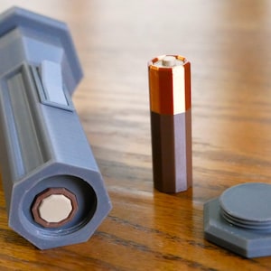 Puede incluir: Una linterna de pl&aacute;stico gris con forma hexagonal y un compartimento para bater&iacute;as. La linterna est&aacute; sobre una superficie de madera junto a una bater&iacute;a marr&oacute;n y una tapa de pl&aacute;stico gris.