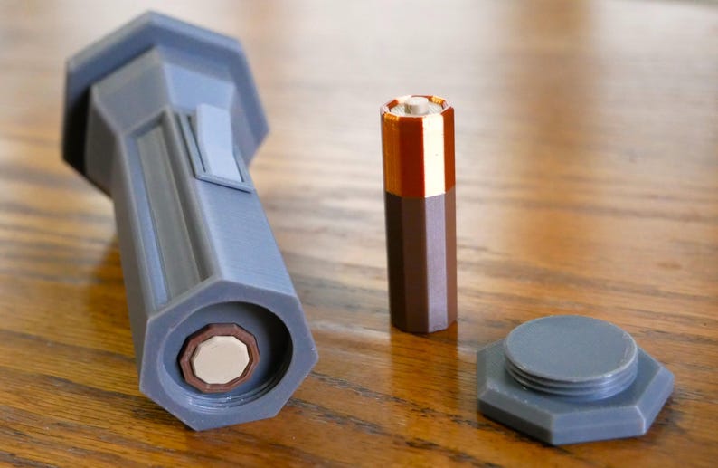 Puede incluir: Una linterna de pl&aacute;stico gris con forma hexagonal y un compartimento para bater&iacute;as. La linterna est&aacute; sobre una superficie de madera junto a una bater&iacute;a marr&oacute;n y una tapa de pl&aacute;stico gris.