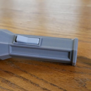 Puede incluir: Una linterna de pl&aacute;stico gris con un interruptor blanco y una bater&iacute;a marr&oacute;n. La linterna tiene una forma hexagonal.