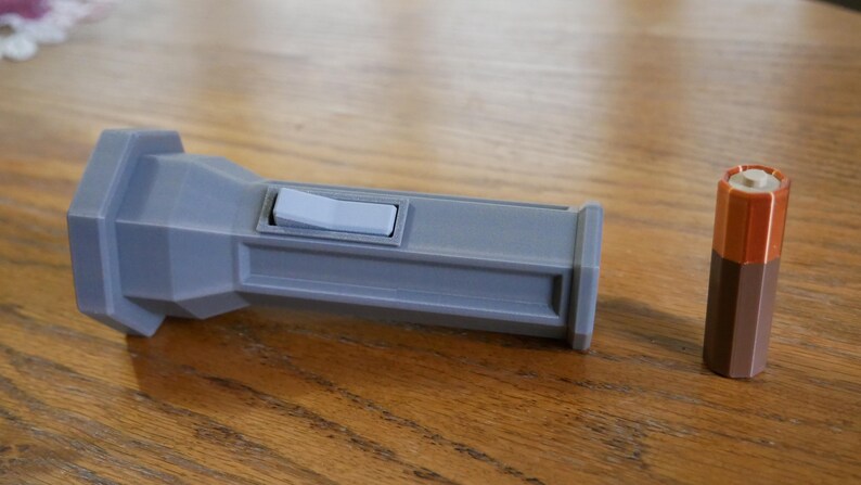 Puede incluir: Una linterna de pl&aacute;stico gris con un interruptor blanco y una bater&iacute;a marr&oacute;n. La linterna tiene una forma hexagonal.