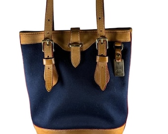May include: A navy blue canvas tote bag with brown leather trim and straps. The bag has two buckles on the front and a small leather tag with a logo.