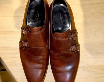 May include: A pair of brown leather double monk strap dress shoes with gold buckles. The shoes have a pointed toe and a polished finish.