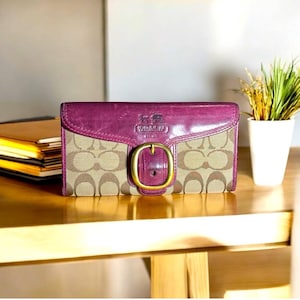 Könnte beinhalten: Eine braune und beige Coach-Geldbörse mit einer großen goldenen Schnalle. Die Geldbörse hat eine violette Lederklappe mit dem Coach-Logo.