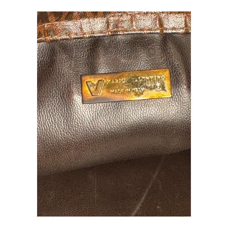 May include: Close-up of a gold-colored leather tag with the text "Mario Valentino Made in Italy" on a brown leather background.