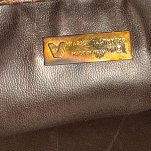 May include: Close-up of a brown leather purse with a gold metal tag that reads "Mario Valentino Made in Italy".