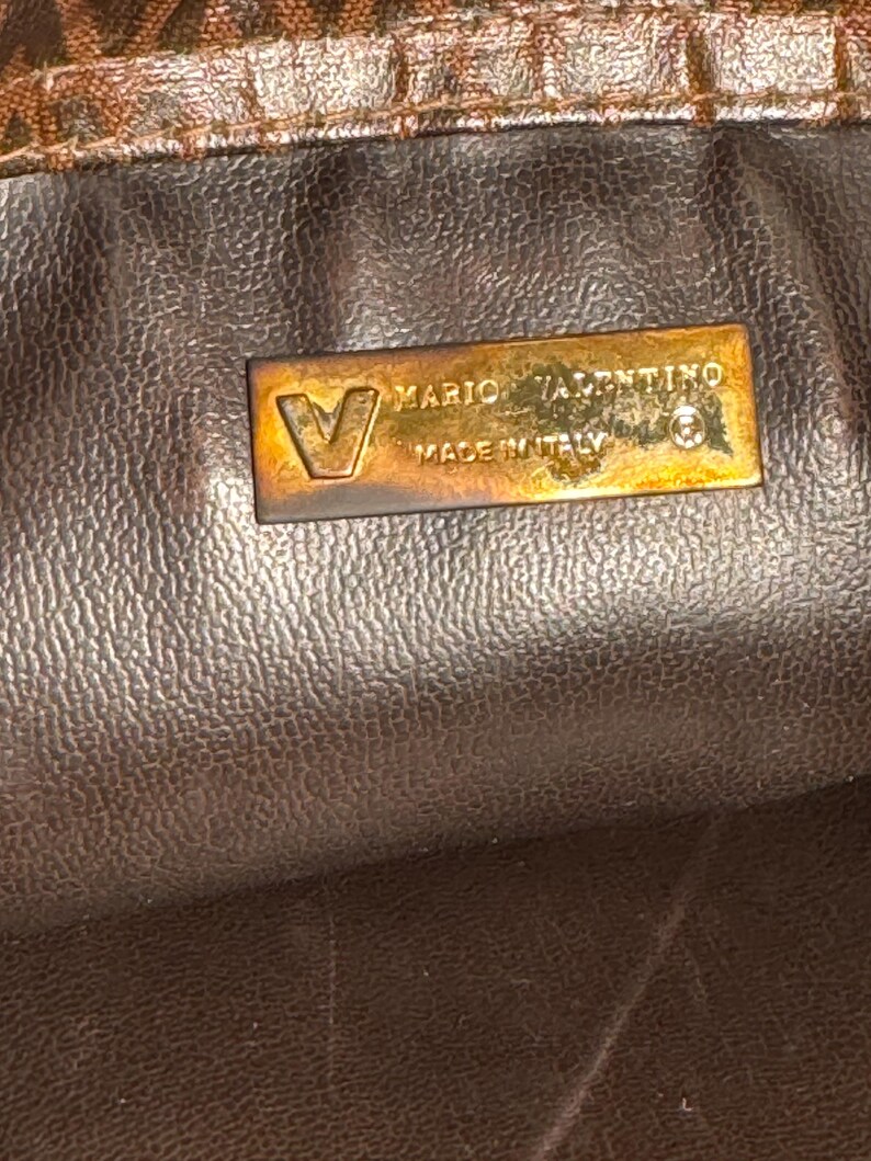 May include: Close-up of a brown leather purse with a gold metal tag that reads "Mario Valentino Made in Italy".