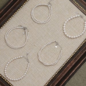 Könnte beinhalten: Fünf Perlenarmbänder, präsentiert auf einer beigen Leinenoberfläche in einem dekorativen braunen Rahmen. Die Armbänder variieren im Design, einschließlich einzelner und doppelter Stränge aus kleinen, weißen Perlen. Jedes Armband hat einen silbernen Verschluss.