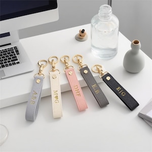 May include: Five personalised keyrings in grey, cream, pink, dark grey, and black. Each keyring has gold-coloured hardware and is engraved with initials. The keyrings are arranged on a white surface.