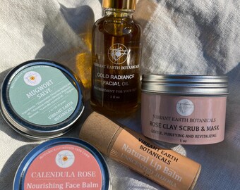 May include: A set of four natural skincare products from Vibrant Earth Botanicals. The products include a mugwort salve, a calendula rose nourishing face balm, a gold radiance facial oil, and a rose clay scrub and mask. The products are packaged in small metal tins and a cardboard tube.