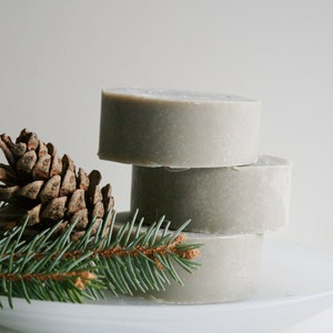 May include: A stack of four round, light gray soaps on a white plate. A brown pine cone and a sprig of green pine needles are arranged on the plate, adding a natural, earthy aesthetic. The soaps appear handmade.